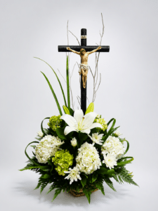 Tributo de Serenidad elegante arreglo fúnebre elaborado con lirios y hortensias, acompañado de helecho cuero, liriope y un Cristo sobre una base de bejuco. Una composición sobria para expresar respeto, condolencias y acompañamiento.
